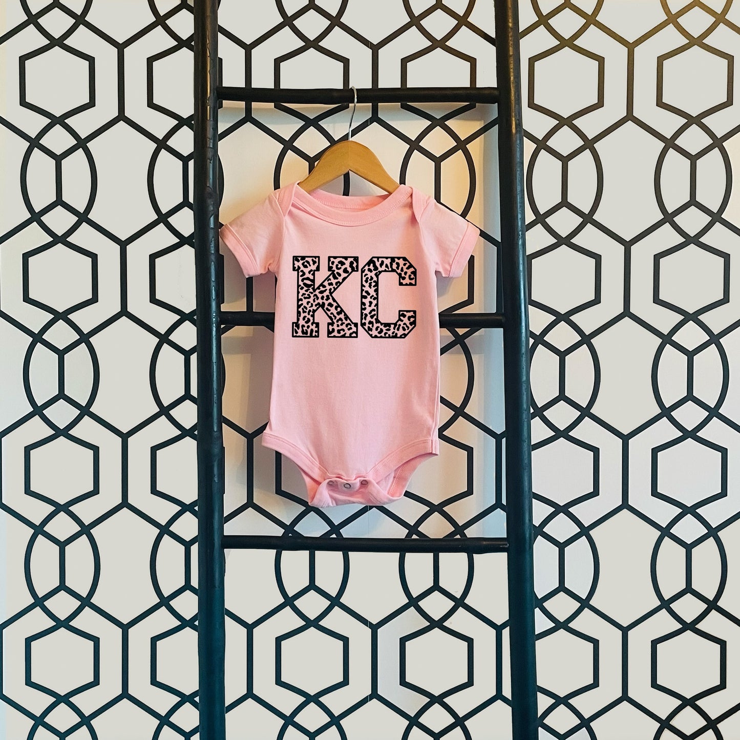 a baby's pink bodysuit hanging on a wall with the letters KC in leopard print