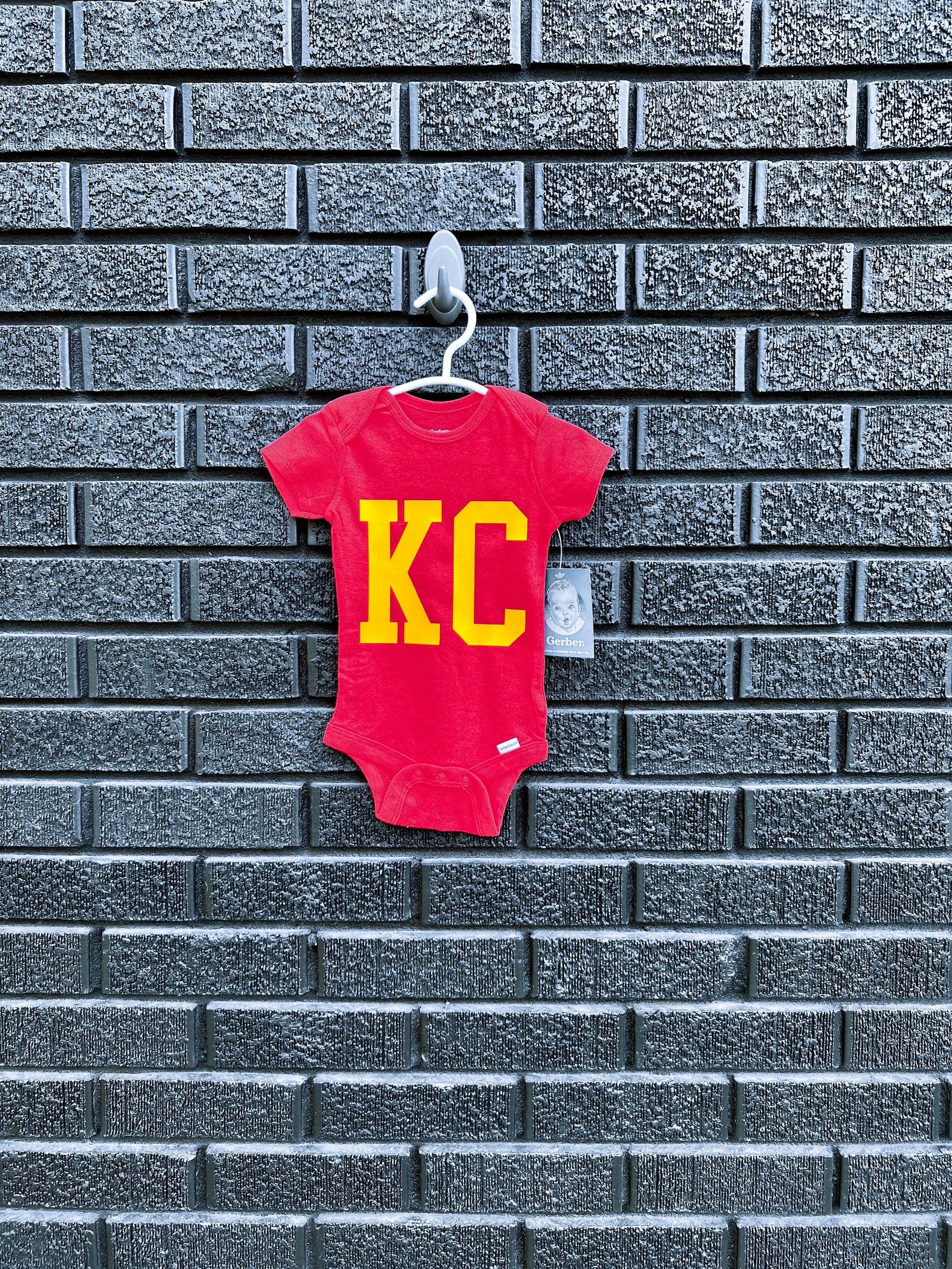 a red onesie with KC hanging on a brick wall