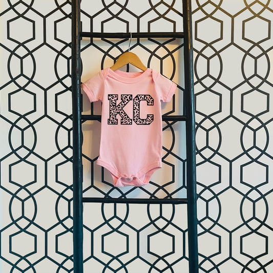 a baby&#39;s pink bodysuit hanging on a wall with the letters KC in leopard print